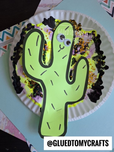 A green paper cactus with googly eyes is glued onto a painted paper plate featuring yellow, black, and purple paint splat designs. The background includes chevron-patterned paper, capturing a playful desert vibe. Text reads @GLUEDTOMYCRAFTS.
