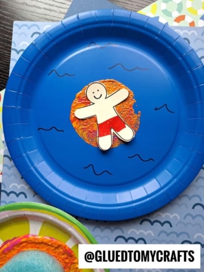 A blue paper plate displays a smiling paper figure in red shorts, floating on a Pool Noodle Stamped Inflatable Raft—a textured orange and yellow circle. The background features colorful patterned paper. @GLUEDTOMYCRAFTS is labeled at the bottom.