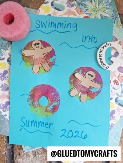 A blue paper with Swimming into Summer 2026 written on it features three paper gingerbread figures in colorful circles, resembling Pool Noodle Stamped Inflatable Rafts. A small pool noodle and a decorative ring are nearby. @gluedtomycrafts is at the bottom.