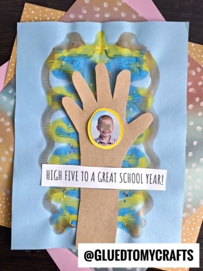 A handmade waving goodbye craft card features a brown paper hand with a childâs photo on the palm, colorful paint patterns as the background, and the text âHigh five to a great school year!â Polka dot paper is layered behind.