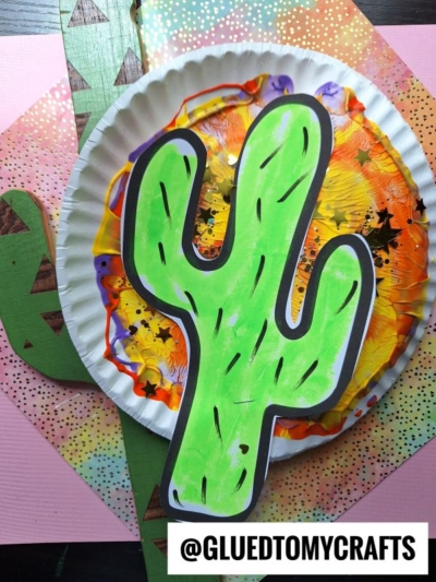 A colorful craft featuring a bright green cactus cutout glued to a painted paper plate with paint splat accents, set on a pink and green patterned background. The handle @GLUEDTOMYCRAFTS appears at the bottom, evoking a playful desert vibe.
