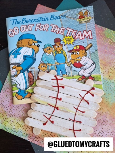 A Berenstain Bears book titled Go Out for the Team sits on colorful paper. In front is a popsicle stick baseball craft laced with red yarn, bringing a sporty touch to the scene. A label reads @GLUEDTOMYCRAFTS.