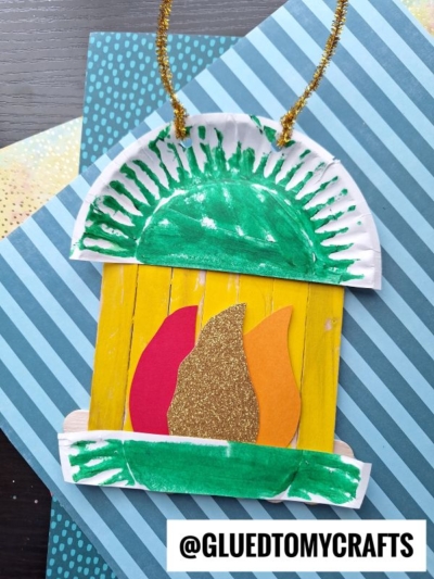 A colorful craft fireplace made from a painted paper plate, popsicle sticks, and cut-out paper flames, with a gold pipe cleaner handle—just like a playful camping lantern—displayed on patterned paper.