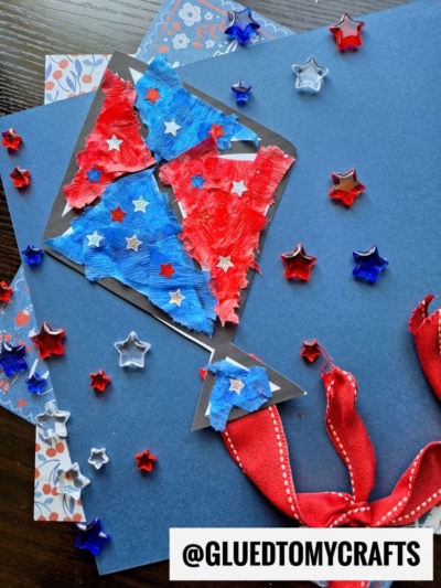 A handmade torn paper kite craft on blue paper, decorated with red and blue crepe paper, star-shaped sequins, and a red ribbon tail. The handle @GLUEDTOMYCRAFTS is visible at the bottom.