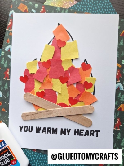 A torn paper campfire art piece features red, orange, and yellow paper flames with small red hearts above two craft stick logs. Below, “YOU WARM MY HEART” is written. A glue bottle and spare paper sit nearby.