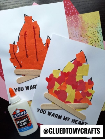 Two torn paper campfire crafts made with colored paper, craft sticks, and glue sit on white sheets labeled You warm my heart. A bottle of Elmer’s glue and colorful background papers are also visible.