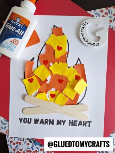 A kids’ craft project showing a torn paper campfire made with orange and yellow pieces, red heart stickers, craft sticks, and glue on a sheet labeled “You warm my heart.” A @gluedtomycrafts label appears at the bottom.