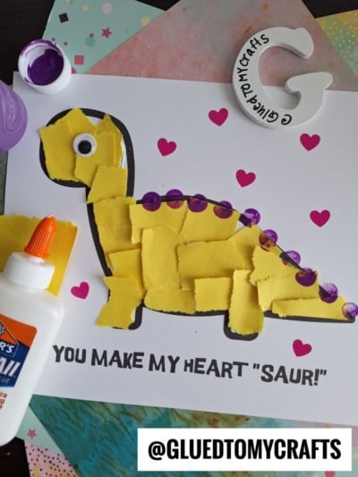 A torn paper dinosaur made of yellow paper pieces, a googly eye, and purple dots on its back, surrounded by pink hearts. Text below reads “YOU MAKE MY HEART ‘SAUR!’” with a glue bottle and paper sheets nearby.