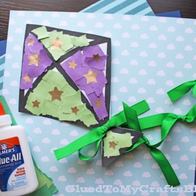 A handmade torn paper kite craft with purple and green pieces and gold stars is glued onto a black outline, displayed on cloud-patterned paper with a bottle of glue and green ribbon bows beside it.