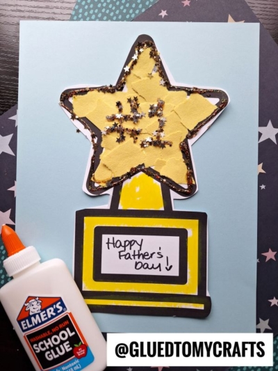 A handmade Fathers Day card shaped like a yellow star trophy with #1 Dad written on it, decorated with glitter and torn paper trophy accents. Below, a plaque reads Happy Fathers Day! Elmers glue sits beside the card; star patterns fill the background.
