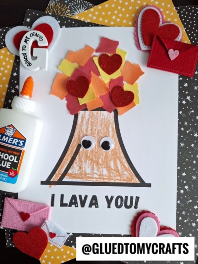 A torn paper volcano made with red, orange, and yellow scraps for lava, plus googly eyes and red hearts, sits on a white page labeled I LAVA YOU! Surrounded by a glue bottle, felt hearts, mini envelopes, and craft materials.