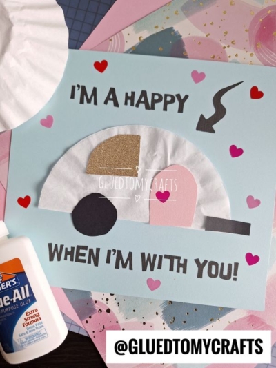 A handmade Valentine’s card with a coffee filter camper, hearts, and the text IM A HAPPY CAMPER WHEN IM WITH YOU! on a blue background. Glue and colorful papers surround the card. Instagram: @GLUEDTOMYCRAFTS.