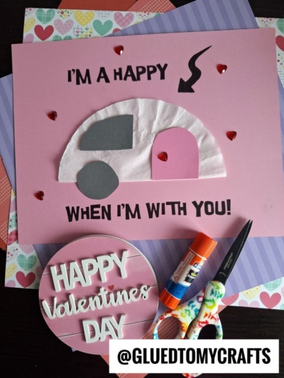 A handmade Valentines Day card features a white and gray coffee filter camper with a pink door on a pink background, heart-shaped gems, and the text I’m a happy camper when I’m with you! Craft supplies are nearby.