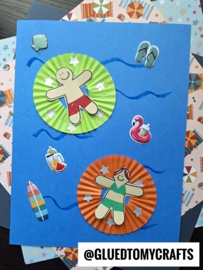 A craft project on blue paper features two paper swimmers on cupcake liner pool floaties, with summer-themed stickers like flip-flops, a drink, and surfboard. Background papers have beach patterns. @GLUEDTOMYCRAFTS is in the corner.