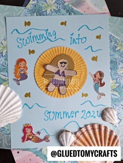 A handmade card with “Swimming into Summer 2024” in blue, decorated with mermaid stickers, shells, and layered paper. A cupcake-liner pool floatie sunshine features a sunglasses-wearing cutout. Background includes patterned papers.