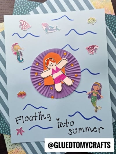 A paper craft features a smiling girl in a swimsuit relaxing on a cupcake liner pool floatie, surrounded by ocean stickers like fish, seahorse, mermaid, and wavy blue lines. Text reads Floating into summer. @GLUEDTOMYCRAFTS at the bottom.