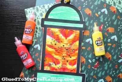 A paint splat camping lantern craft with a wavy, colorful center and bold black outline sits on patterned paper, surrounded by bottles of orange, yellow, and red puffy paint.