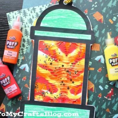 A paint splat camping lantern craft with a wavy, colorful center and bold black outline sits on patterned paper, surrounded by bottles of orange, yellow, and red puffy paint.