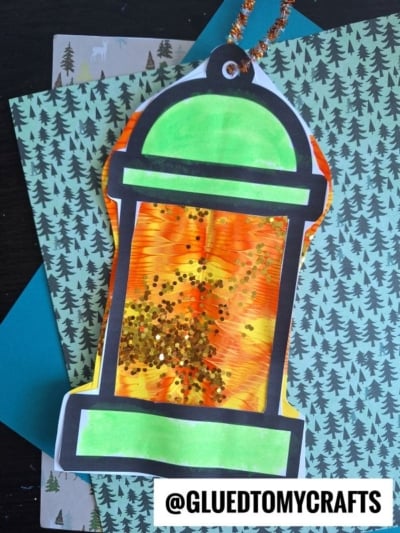 A colorful paint splat camping lantern craft with orange, yellow, and green is decorated with glitter and bordered in black, resting on pine tree-patterned paper. @GLUEDTOMYCRAFTS is printed at the bottom.