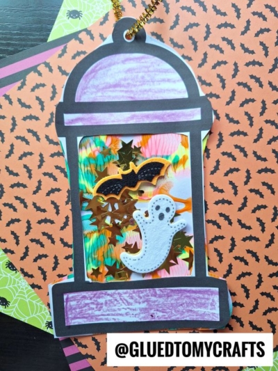 A Halloween-themed paint splat camping lantern craft featuring a bat, gold stars, and a ghost on colorful paper, set against patterned orange and green backgrounds. The text @GLUEDTOMYCRAFTS appears at the bottom.