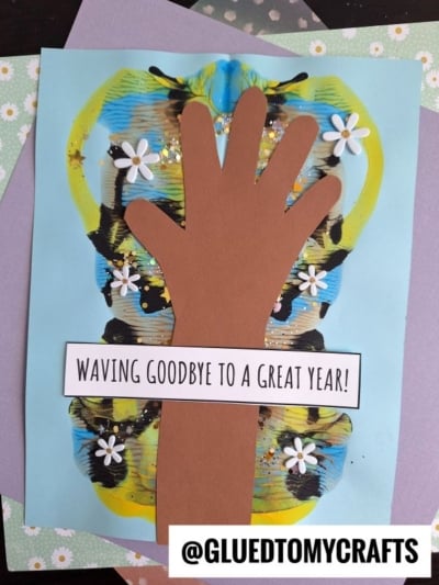A child's brown paper hand is glued on colorful butterfly-shaped paint blobs over blue paper, decorated with white flower accents and glitter. This waving goodbye craft features a label: WAVING GOODBYE TO A GREAT YEAR! @GLUEDTOMYCRAFTS at the bottom.