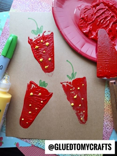 A craft project featuring four red strawberry prints with yellow dot details on brown paper, created using a pie server stamped technique. A red paint-covered paper plate, yellow paint bottle, and green marker are nearby; @GLUEDTOMYCRAFTS is shown at the bottom.