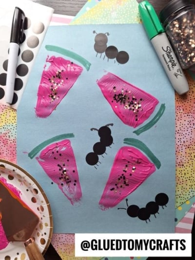 A blue paper displays five watermelon slice paintings with pink paint and gold glitter, two black ant shapes, and a pie server stamped design. Art supplies like Sharpie markers, dot stickers, and glitter are nearby. @GLUEDTOMYCRAFTS is written at the bottom.