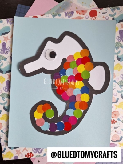 A polka dot seahorse craft made with colorful paper circles and a googly eye is glued onto a light blue background, surrounded by patterned papers. @GLUETOMYCRAFTS is written at the bottom of the image.
