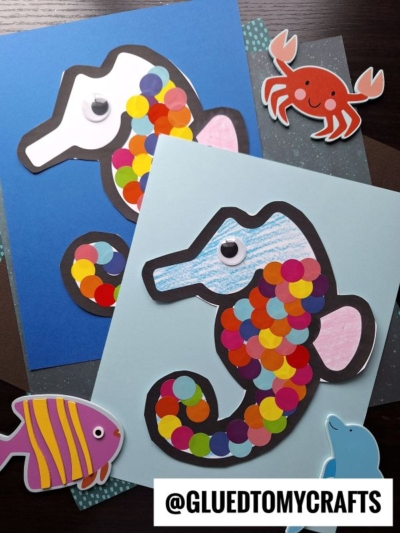 Two colorful polka dot seahorse crafts made from paper, decorated with bright dots and googly eyes, are surrounded by paper cutouts of a crab and a fish. The handle @GLUEDTOMYCRAFTS appears at the bottom.