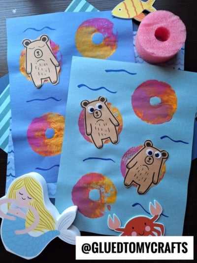 Two blue papers with bear cutouts and colorful painted rings, resembling pool floats in a lazy river bear craft. Surrounding them are a fish, a pool noodle slice, a mermaid, and a crab decoration. @GLUEDTOMYCRAFTS is written at the bottom.