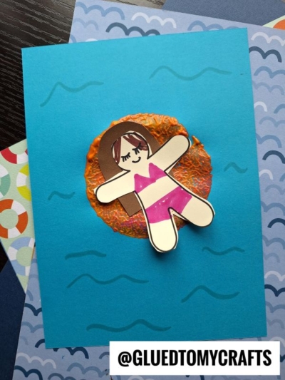 A handmade card features a paper cutout of a girl in a pink swimsuit floating on a textured orange circle, resembling a Pool Noodle Stamped Inflatable Raft, with blue wavy lines for water. The username @GLUEDTOMYCRAFTS appears at the bottom.