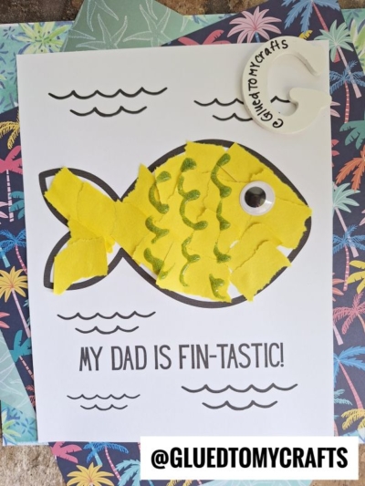 A handmade Father’s Day card featuring a yellow fish made from torn paper, a googly eye, and green squiggles. The text celebrates your fin-tastic dad with “MY DAD IS FIN-TASTIC!” and a tag that says “@GLUEDTOMYCRAFTS.”.