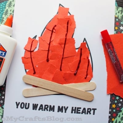 A torn paper campfire craft with red and orange flames, two craft sticks for logs, and “You warm my heart” text on a white background. Glue and a red marker complete this cozy torn paper campfire project.