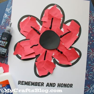A handmade torn paper poppy flower craft made with red paper pieces glued to a black outline, with REMEMBER AND HONOR written below.