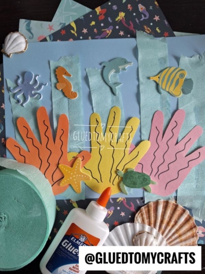 A colorful undersea-themed craft features a handprint coral reef made with paper handprints, foam sea animals, shells, tissue paper, glue, and craft supplies on a blue background. The Instagram handle @GLUEDTOMYCRAFTS appears at the bottom.