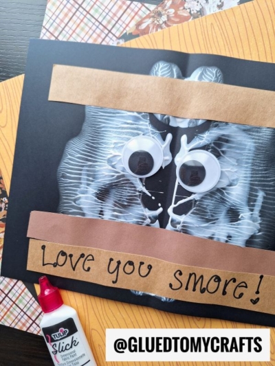 Handmade card with an X-ray image, two large googly eyes, and brown paper strips that say Love you S’more! A playful paint splat adds extra charm, while a bottle of pink craft glue and patterned papers are nearby. @GLUEDTOMYCRAFTS label at the bottom.