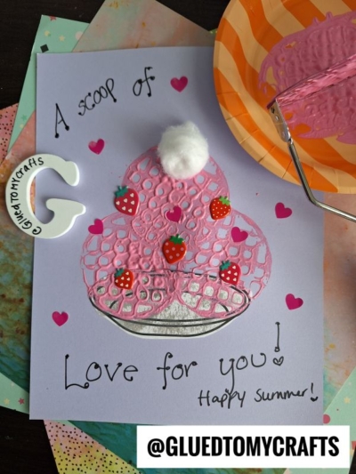 A handmade card with a pink potato masher stamped ice cream sundae topped with a white pom-pom and small strawberry stickers. The text reads, “A scoop of Love for you! Happy Summer!” Art supplies are around the card.