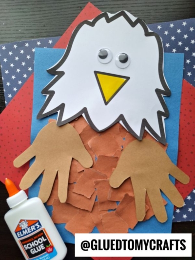 A torn paper eagle craft made from paper, featuring a white head, yellow beak, googly eyes, handprint wings, and a body of brown paper pieces. An Elmers glue bottle and “@GLUEDTOMYCRAFTS” label are visible.