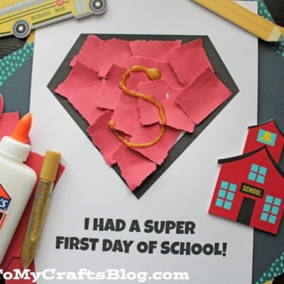 A school-themed craft features a torn paper superhero S emblem, resembling Superman’s logo, on a worksheet saying "I HAD A SUPER FIRST DAY OF SCHOOL!" Surrounded by glue, crayons, and fun school cutouts.