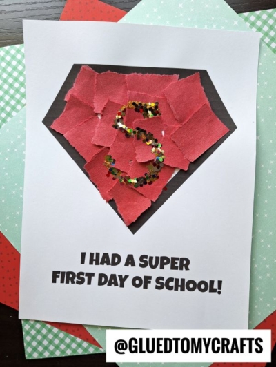 A kids’ craft features a torn paper superhero shield—black with red paper pieces glued inside and a sparkly S in the center. Below it reads, I HAD A SUPER FIRST DAY OF SCHOOL! and @GLUEDTOMYCRAFTS.