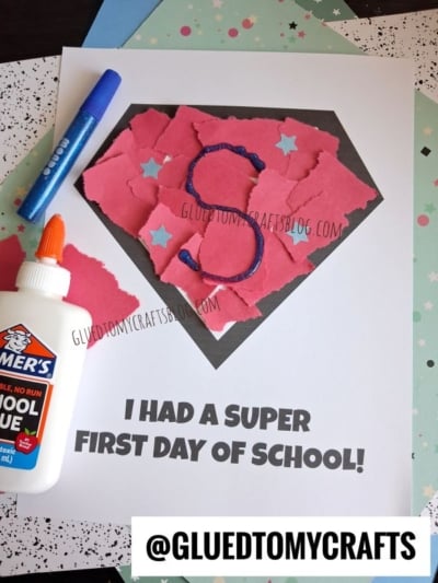 Torn pink paper forms a torn paper superhero logo, reminiscent of Superman, with a blue S on a worksheet that says I HAD A SUPER FIRST DAY OF SCHOOL! Nearby are glue, a blue glitter pen, and patterned paper.