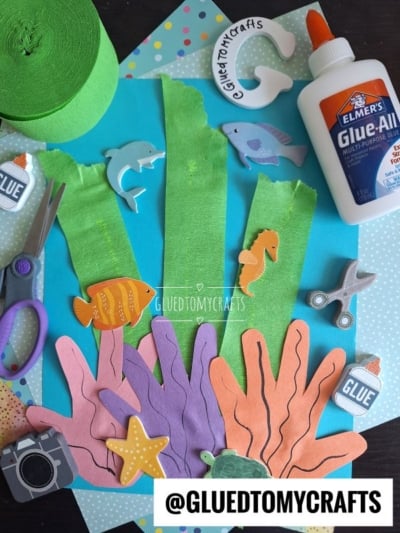 A flat lay of ocean-themed craft supplies, perfect for making a handprint coral reef—featuring green crepe paper strips, sea animal foam cutouts, scissors, glue, and blue polka dot paper. The handle @GLUEDTOMYCRAFTS is displayed at the bottom.