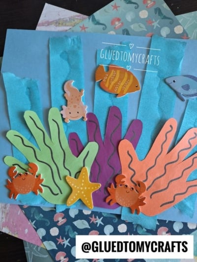 Colorful, hand-shaped paper cutouts create a vibrant handprint coral reef with seaweed, crabs, a starfish, a fish, and a dolphin on a blue background. The handle @gluedtomycrafts is visible.