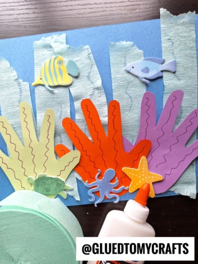 A colorful ocean-themed craft featuring handprint coral reef cutouts, crepe paper seaweed, foam sea animals, a glue bottle, and a roll of green crepe paper on a blue background. @GLUEDTOMYCRAFTS is written at the bottom.