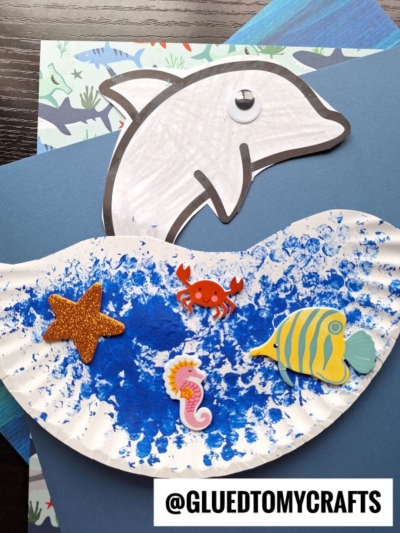 A paper plate dolphin cutout and ocean-themed stickers, like a starfish, crab, seahorse, and fish, decorate a blue and white painted wave. The background features patterned paper with sea animals. @GLUEDTOMYCRAFTS is displayed at the bottom.