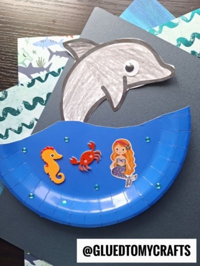 A paper plate dolphin craft featuring a gray dolphin jumping from blue water, decorated with seahorse, crab, and mermaid stickers on ocean-themed paper. @GLUEDTOMYCRAFTS is at the bottom.
