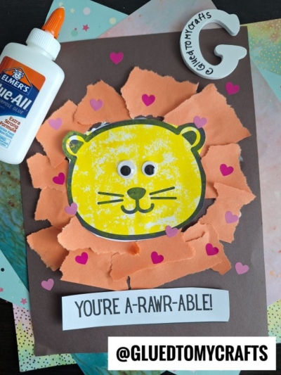 A handmade torn paper lion card with a yellow face, googly eyes, and an orange mane on brown paper. Pink heart stickers decorate it. Nearby are a glue bottle, craft supplies, and a note saying “YOU’RE A-RAWR-ABLE!”.