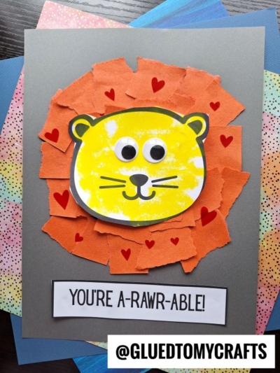 A handmade card showcases a torn paper lion with a smiling yellow face and googly eyes, surrounded by red heart stickers. The message YOURE A-RAWR-ABLE! appears at the bottom, making it extra charming.