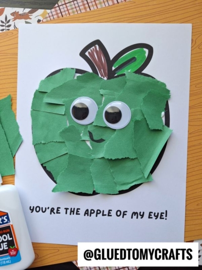 A torn paper apple craft features a green apple with large googly eyes on white paper. Text reads “YOU’RE THE APPLE OF MY EYE!” Glue and green paper pieces are visible nearby, highlighting the fun of creating this adorable torn paper apple project.