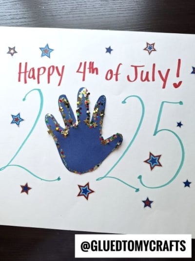 A handmade 4th of July keepsake features "Happy 4th of July!" in red, "2025" in teal, a blue handprint with star-shaped confetti, and star stickers on white paper. The handle @GLUEDTOMYCRAFTS is at the bottom.